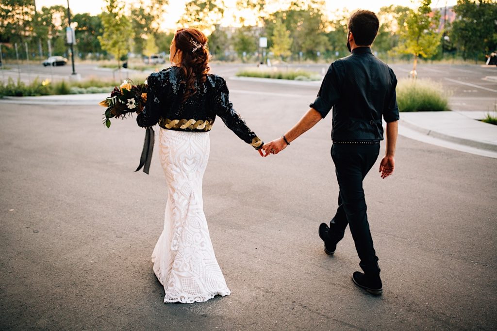 A newlywed couple walk hand in hand away from the camera in a parking lot wearing their wedding attire.