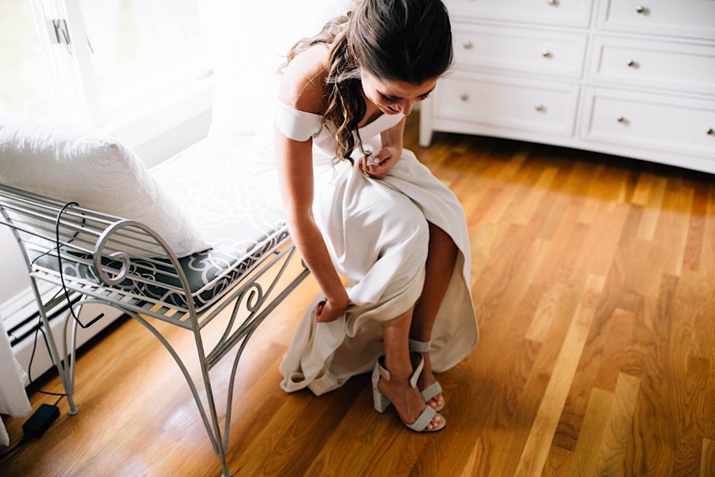 A bride in her wedding dress puts on her dress shoes while seated.