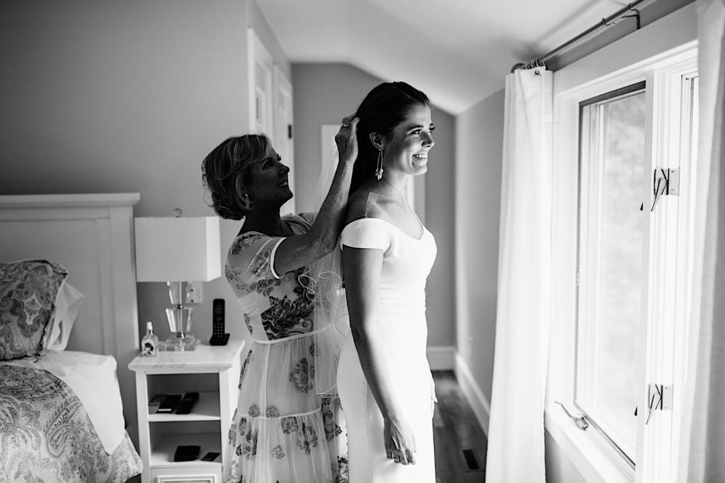 Black and white photo of a bride in her wedding dress smiling and looking out the window as her mother stands behind her smiling and adjusting her hair.