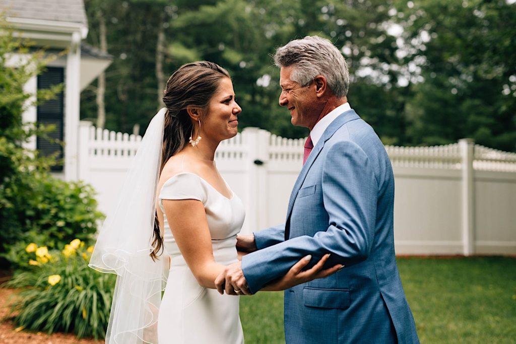 A bride and her father stand outside facing one another and smile, they're in their wedding attire and the bride is starting to tear up with emotion.