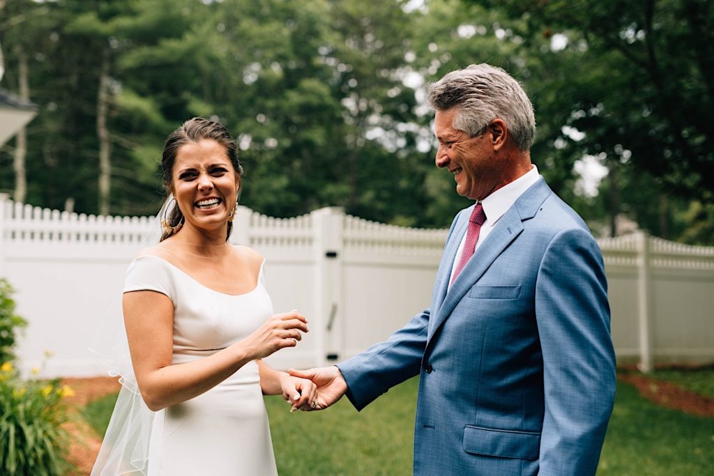 A bride laughs and smiles at the camera as her father smiles at her while holding her hand. The two are standing outside their home in front of a white fence.