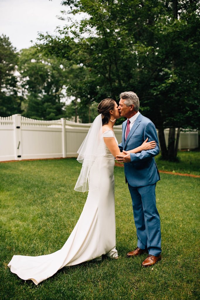 A bride and her father stand outside of their home in their wedding attire and kiss one another on the cheek