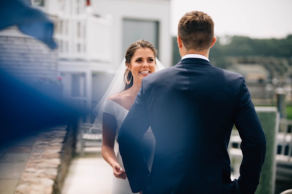 A bride and groom share their first look outside their Cape Cod wedding venue, the groom's back is turned to the camera and the bride is smiling