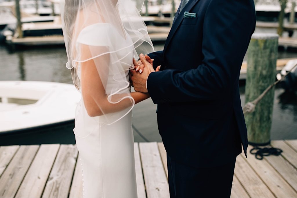A bride and groom hold hands while wearing their wedding attire, there is a boat dock behind them.