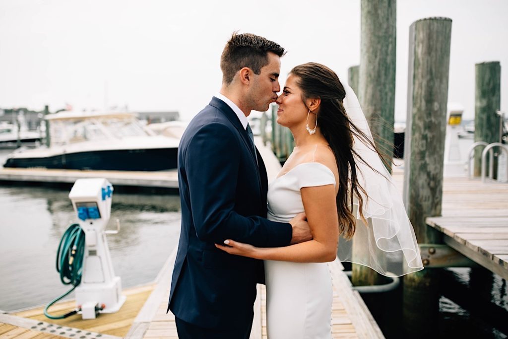 A groom kisses his bride on the nose as she smiles. Both are dressed for their wedding and are standing on a boat dock in Cape Cod