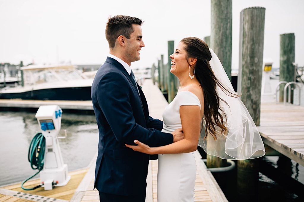 A bride and groom dressed for their wedding face one another and smile as they embrace, they are standing on a boat dock in Cape Cod