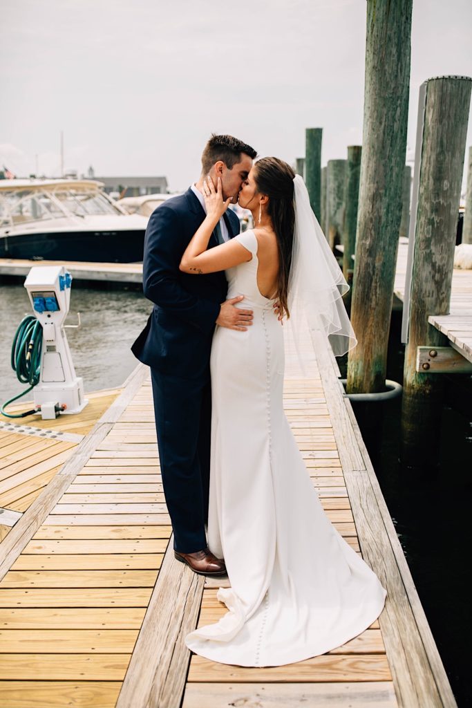 A bride and groom dressed for their wedding kiss one another as they stand on a boat dock in Cape Cod