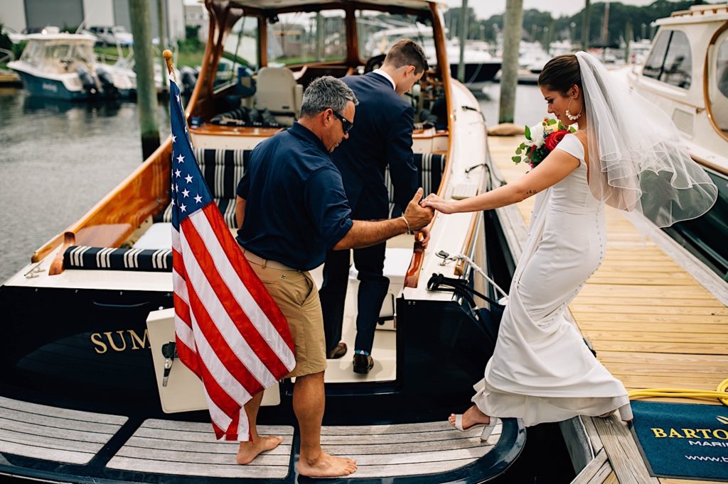 A bride in her wedding dress is helped onto a small docked boat by a man just after he helped her groom aboard.
