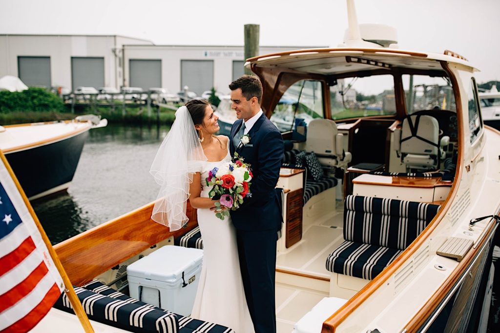 A bride and groom in their wedding attire stand next to one another and smile at each other. They are on a small docked boat in Cape Cod