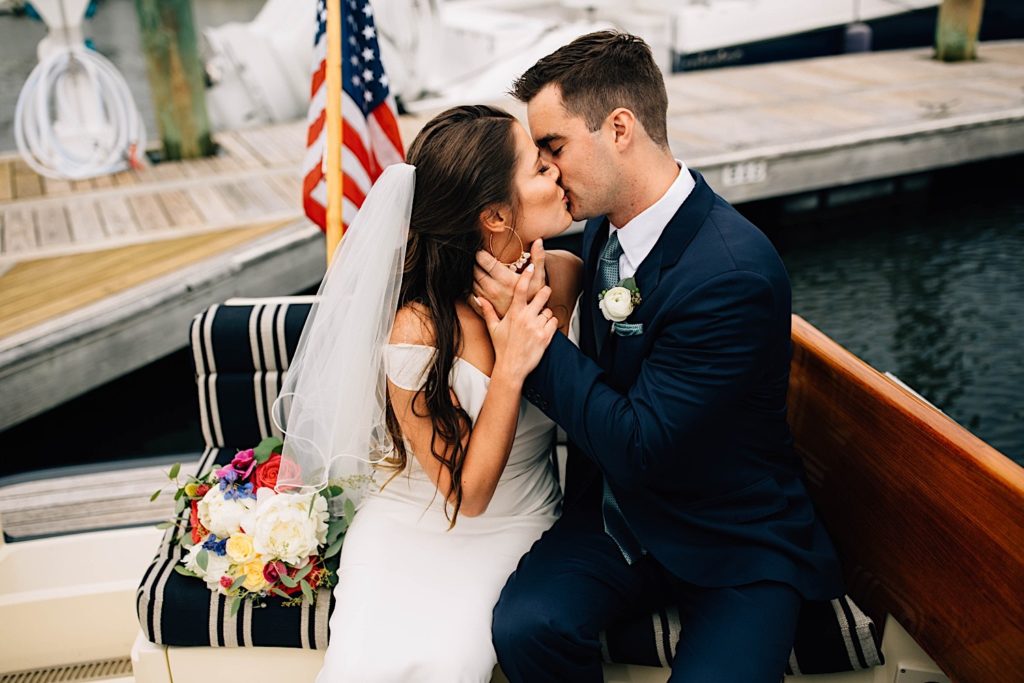 A bride and groom dressed for their wedding sit next to one another and kiss on a docked boat