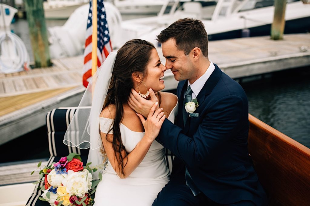 A bride and groom dressed for their wedding sit next to one another and smile on a docked boat