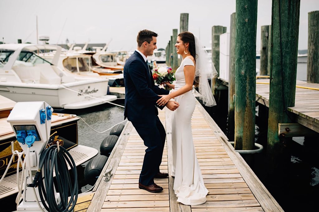 A bride and groom dressed for their wedding stand facing one another and smile as the groom dances, they're standing on a boat dock outside their Cape Cod wedding venue
