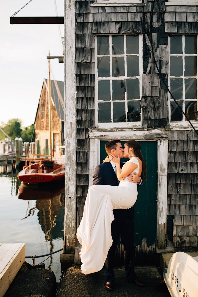 A bride and groom kiss in front of an old building on the water, their in their wedding attire and the groom is carrying the bride.