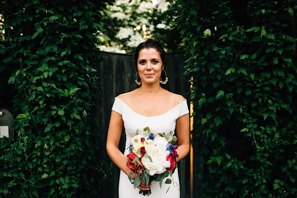 A bride in her wedding dress holds a flower bouquet and gives a soft smile to the camera