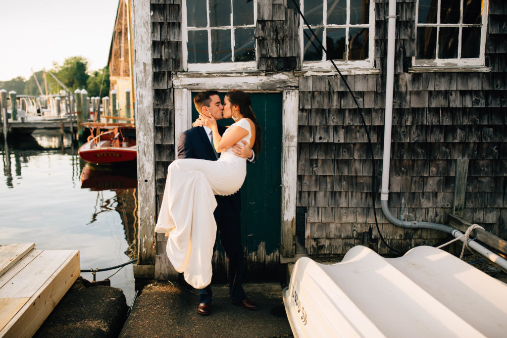 A bride and groom kiss in front of an old building on the water, their in their wedding attire and the groom is carrying the bride.