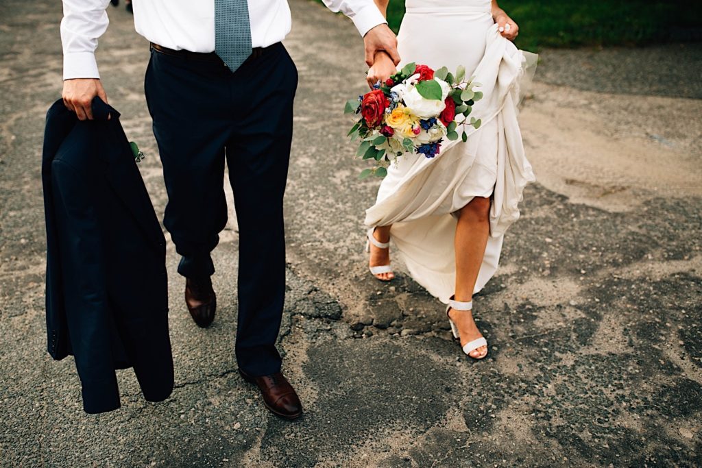 Photo of a bride and groom waist down in their wedding attire walking towards the camera, the groom is holding his suit coat in one hand and his brides arm in the other, the bride is holding a flower bouquet