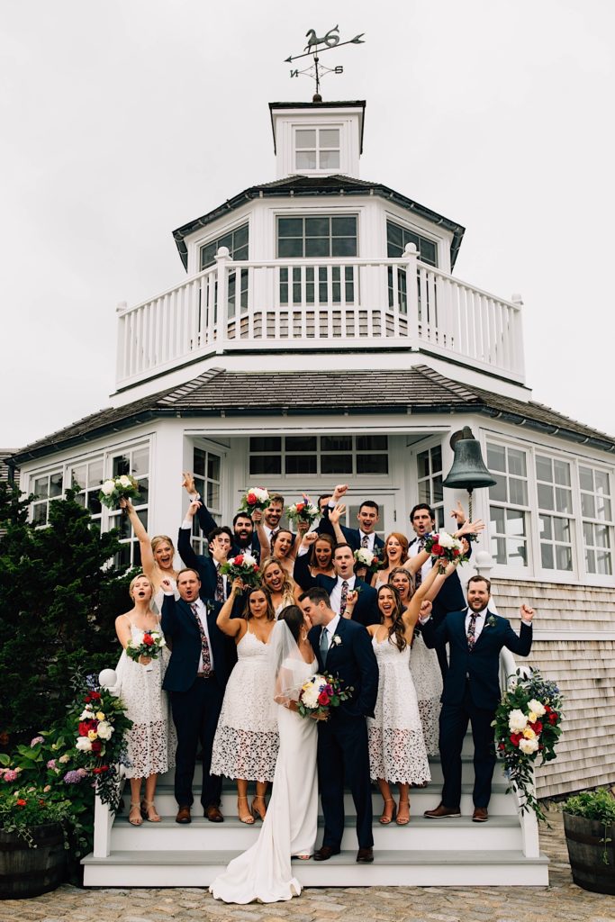 A bride and groom kiss with their wedding parties cheering behind them, the group is standing on the steps leading up to their Cape Cod wedding venue.