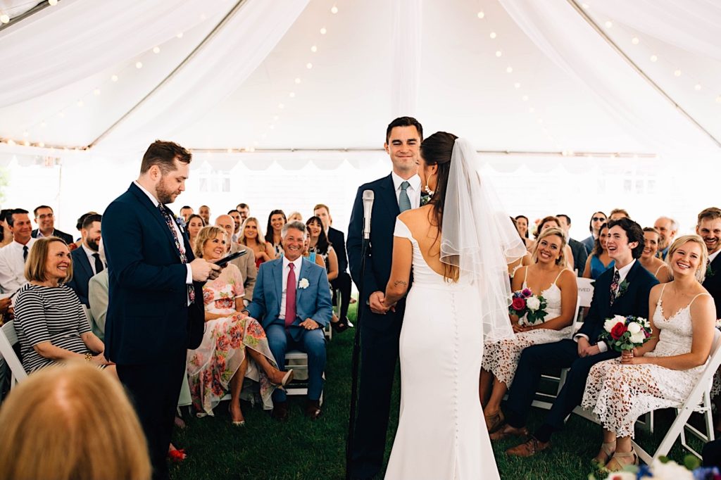 A bride and groom stand holding hands during their wedding ceremony that is under a tent with stringlights, the officiant is speaking while their guests are seated in a circle around them