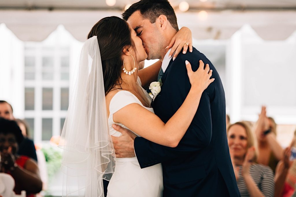 A bride and groom embrace and kiss during their wedding ceremony while guests clap and cheer behind them.