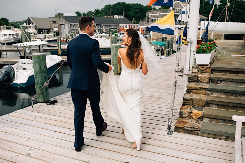 A bride and groom walk away from the camera on a boat dock in Cape Cod after their wedding ceremony. The groom is holding the bride's dress while she walks and smiles at him.