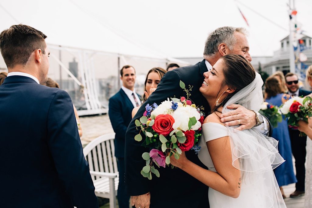 A father hugs his daughter after her wedding ceremony with guests in the background all smiling.