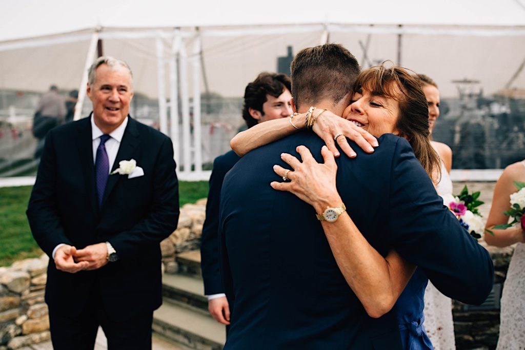 A mother hugs her son after his wedding ceremony while his father stands to the left of them and smiles.