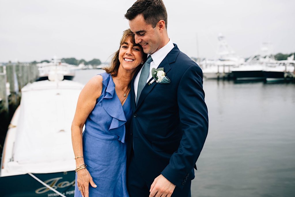 A groom stands next to his mother as they both smile and pose for a picture on a boat dock.