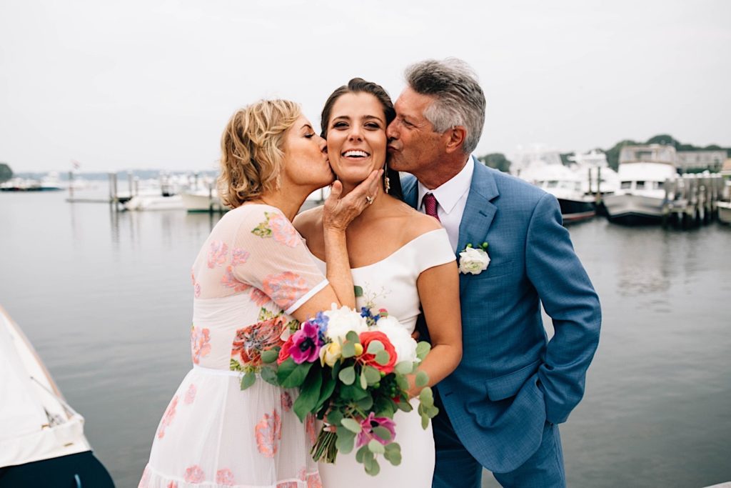 A bride in her wedding dress stands between her mother and father as they both kiss her on the cheek at a boat dock in Cape Cod