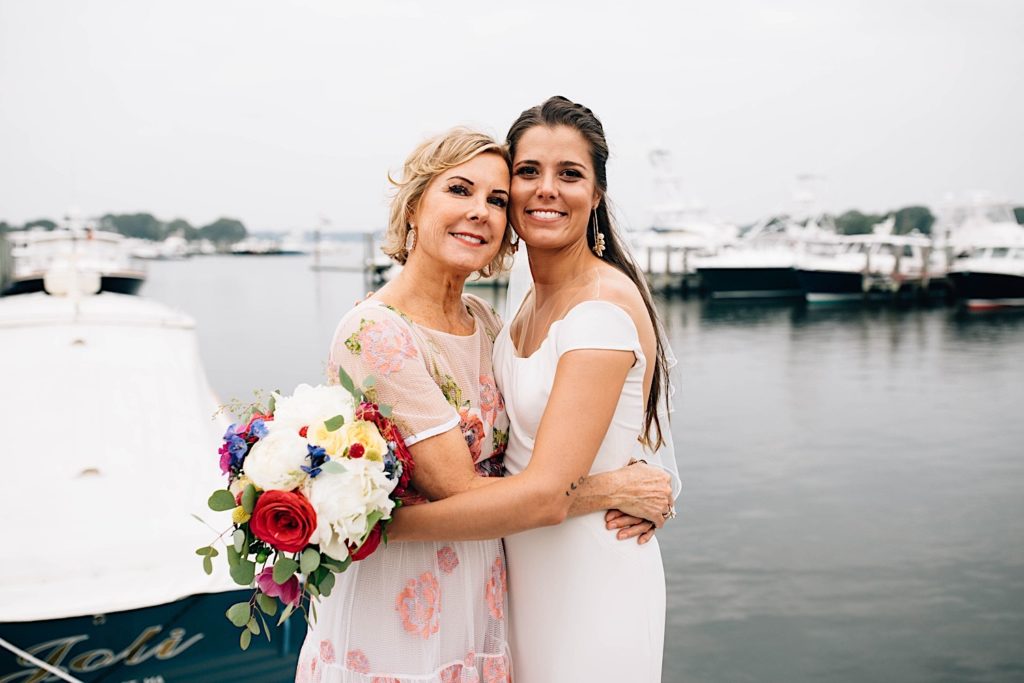 A bride and her mother hug one another and smile at the camera after a wedding ceremony.