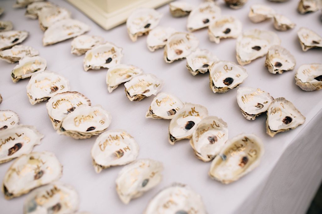 Half oyster shells are laid out on a table with names and numbers written on them, they are for wedding guests to know which table to sit at.