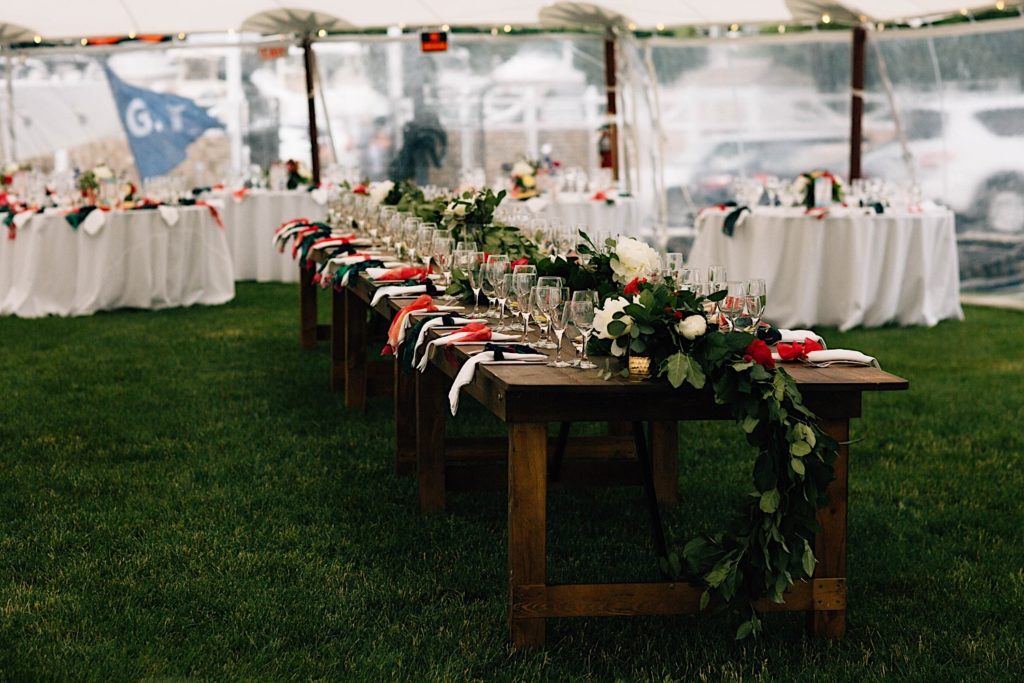 A long wooden table set up for a wedding with wine glasses, flowers and napkins, the table is underneath a tent with smaller circular tables behind it.