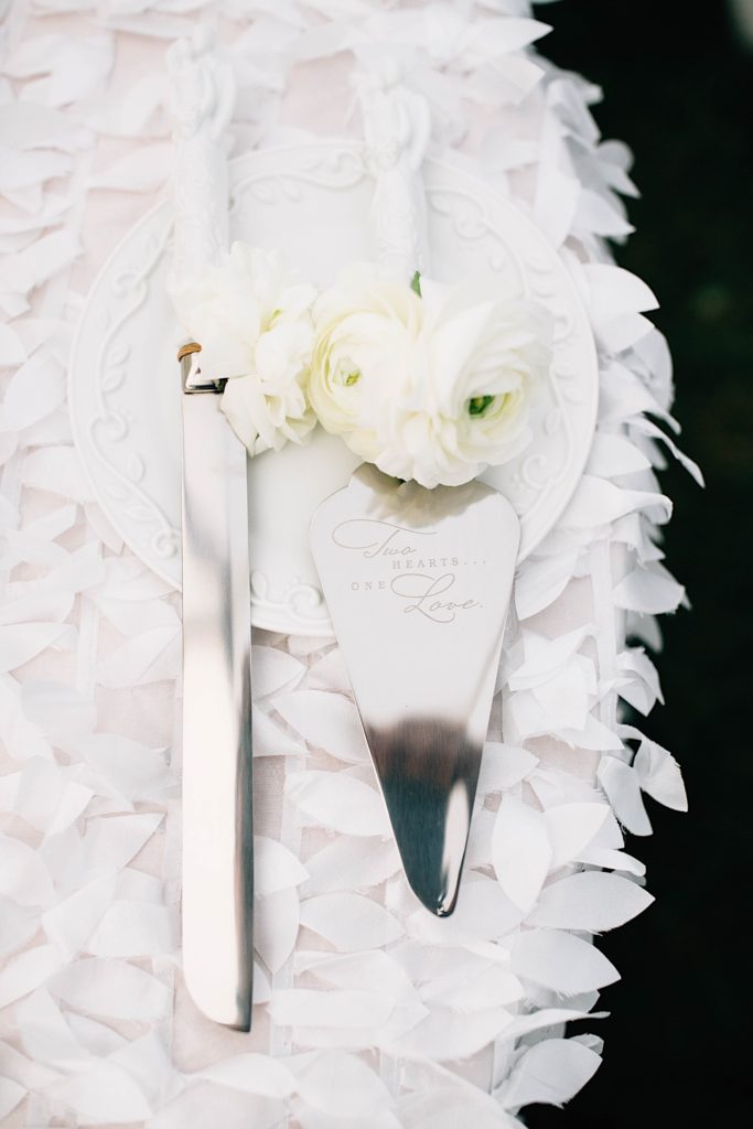 Close up photo of cake knives with white flowers on the handles, the larger knife has "Two hearts...one love" engraved on the blade.