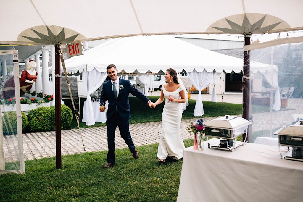 A bride and groom dressed for their wedding walk into their wedding reception tent together while holding hands and smiling.