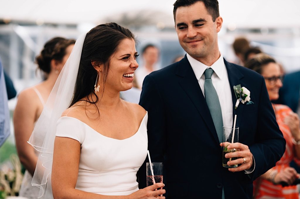 A bride and groom stand side by side in their wedding attire and smile during their wedding reception. The bride is laughing at something out of frame while the groom smiles at her.