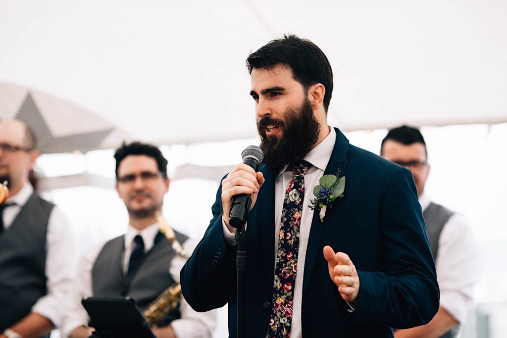 A groomsmen at a wedding reception gives a speech while the live band stands behind him.