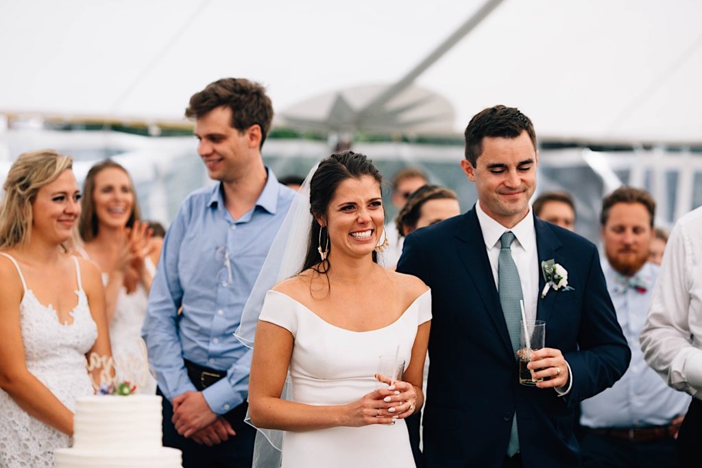 A bride and groom stand next to one another and smile with guests standing behind them during their Cape Cod wedding reception.