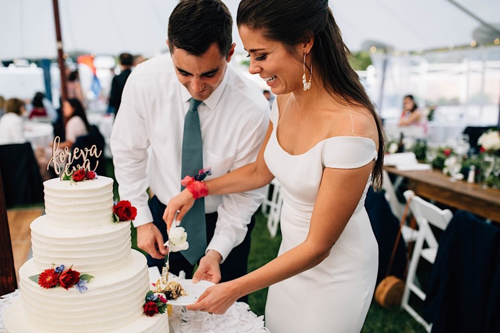 A bride and groom stand together and smile as they cut the cake during their wedding reception.