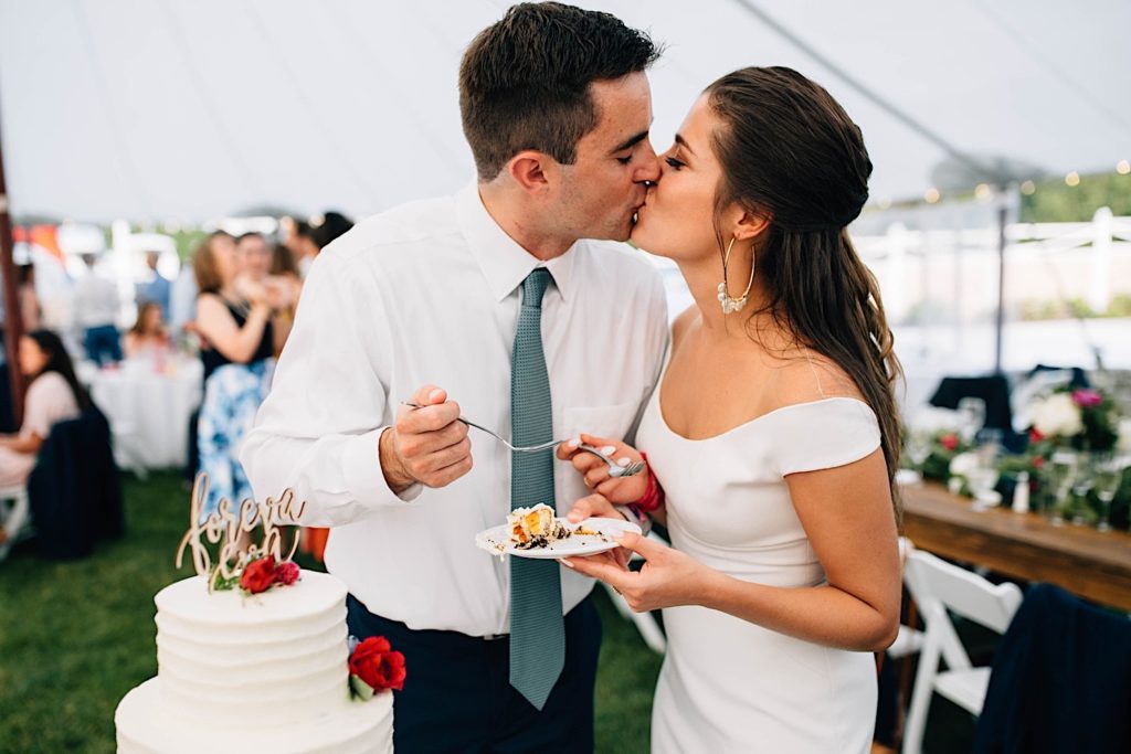 A bride and groom kiss one another during their Cape Cod wedding reception, the bride is holding a plate with a piece of cake on it.