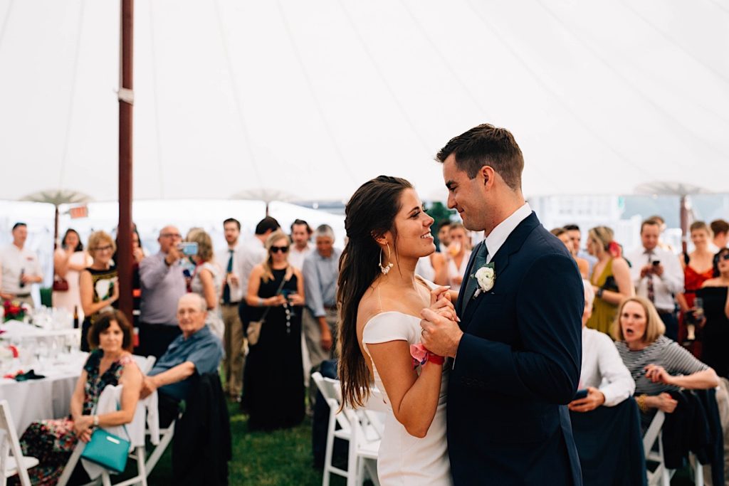 A bride and groom smile at one another during their first dance during their Cape Cod wedding reception underneath a large white tent.