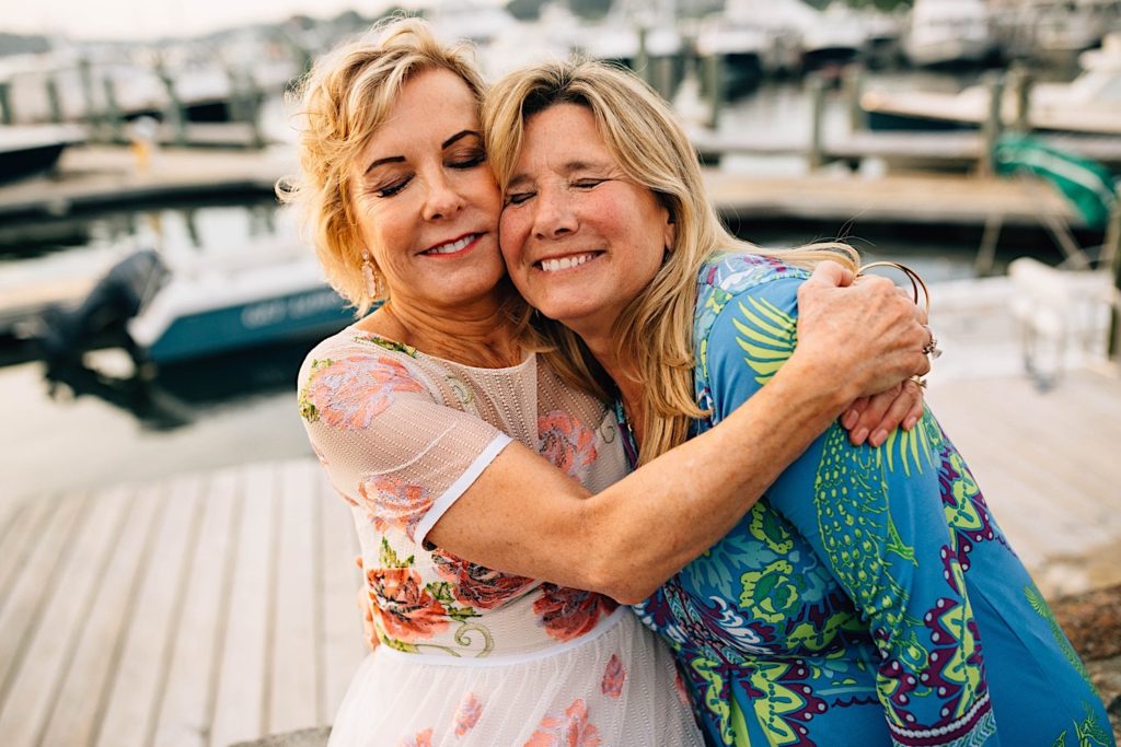 Two women dressed for a wedding reception smile and hug on a boat dock.