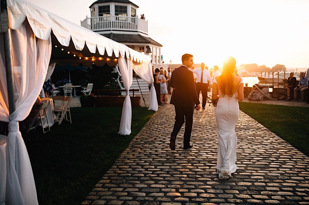 A bride and groom walk away from the camera towards the sunset after their Cape Cod wedding reception.