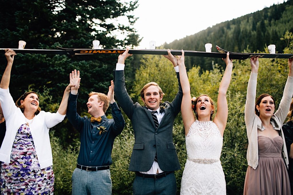 A bride, groom and 3 members of their wedding party stand side by side holding skis above their heads with drinks on the skis. The groom is smiling at the camera while the others laugh