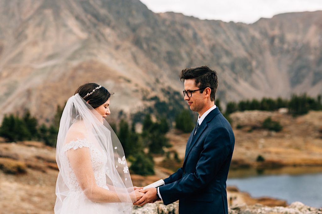 A bride and groom stand facing one another holding hands while wearing their wedding attire with mountains and a lake behind them.