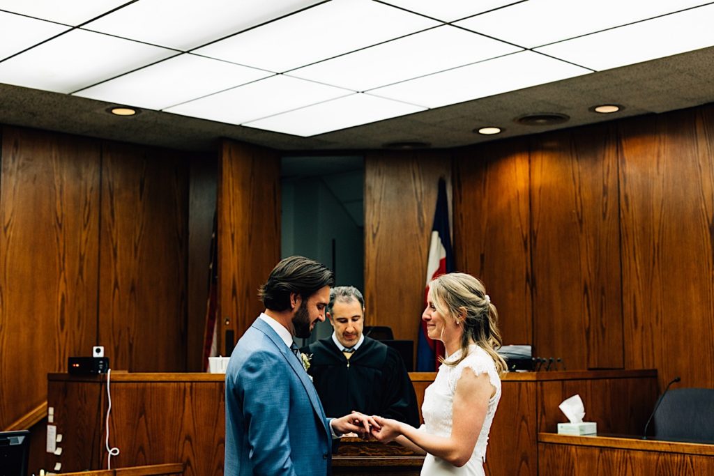 A bride and groom stand facing one another holding hands as they are married in a courtroom.