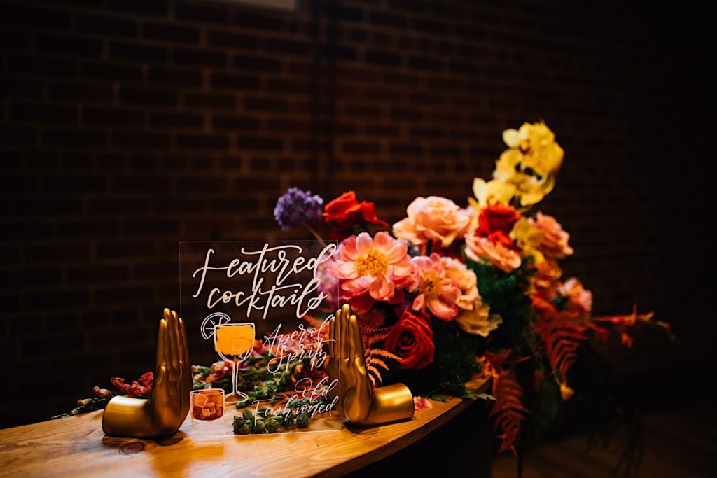 A glass sign with featured cocktails written on it held up by 2 gold hands that are bookends. The sign is on a wooden bench with colorful flowers behind it