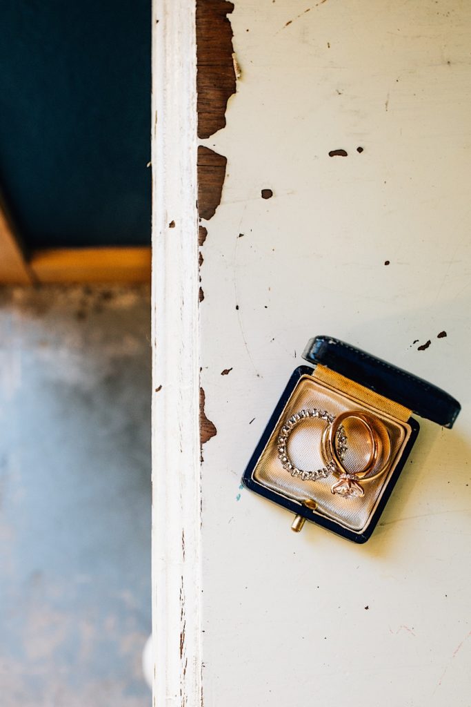 The bride and grooms wedding rings sitting in a black leather ring box on a rustic white dresser with paint chipping to show wood underneath