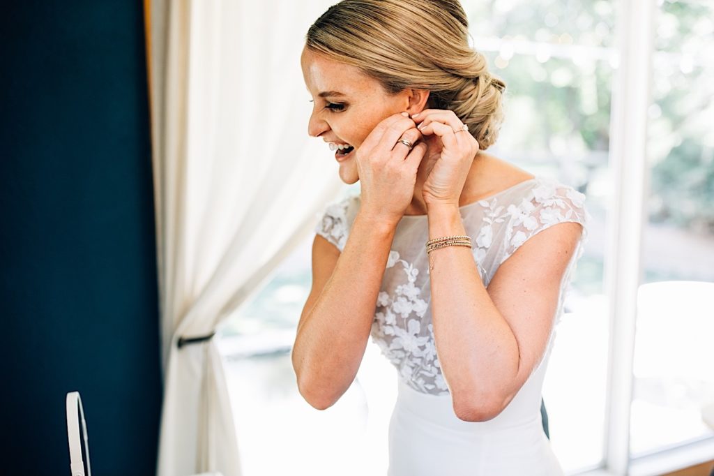 Bride puts on her wedding day earrings while smiling into the mirror.