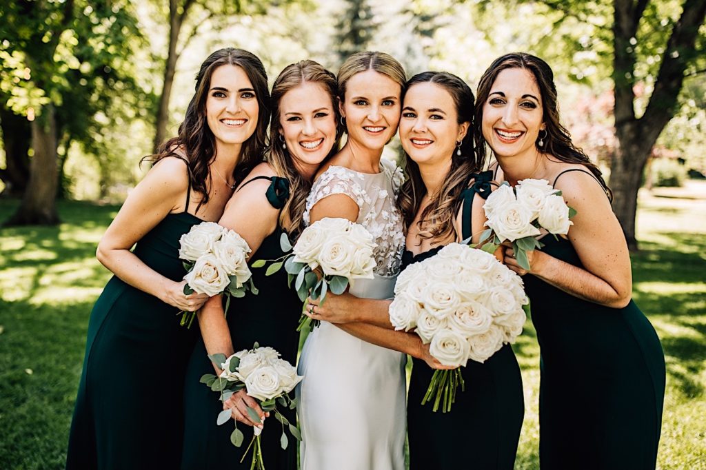 Bride and bridesmaids smile together for a photo.  The bridesmaids wear all black wedding attire and everyone holds bouquets of white roses